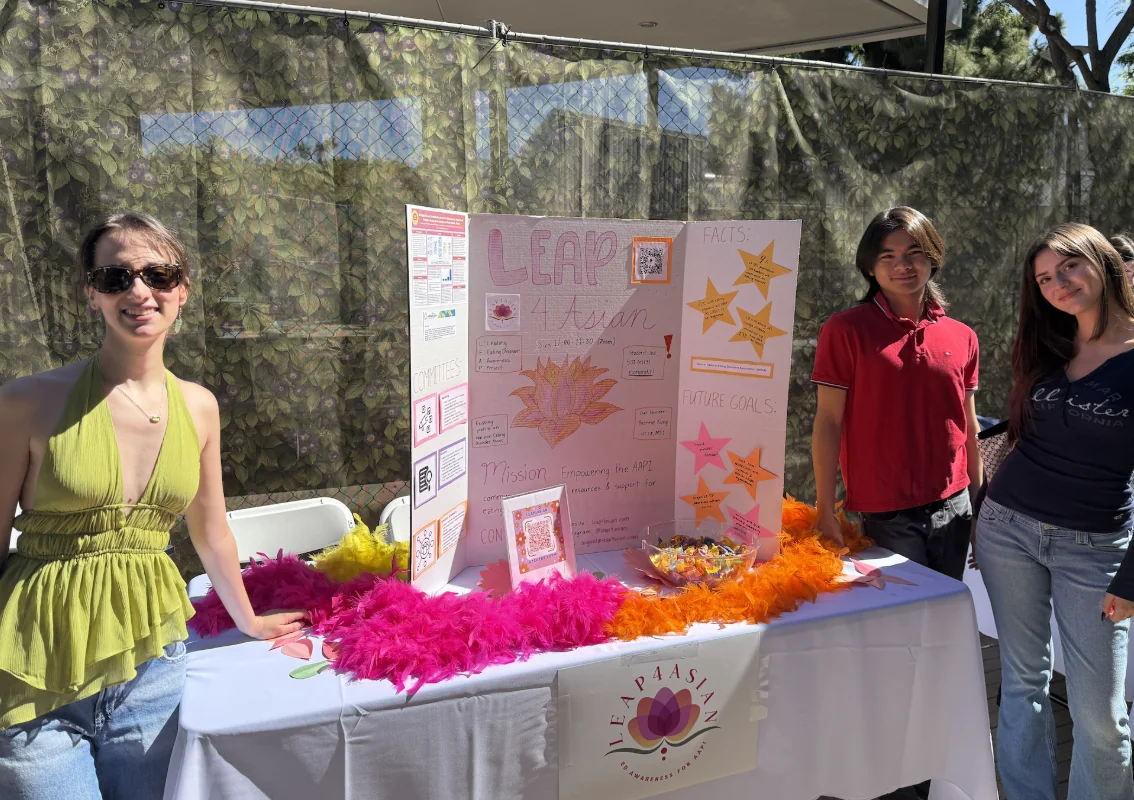Three LEAP4Asian volunteers stand behind an outdoor tabling booth with a white tablecloth, a trifold display board reading LEAP4Asian with committees, facts, and future goals panels, feather boas in pink and orange, a framed QR code sign, and a bowl of candy.